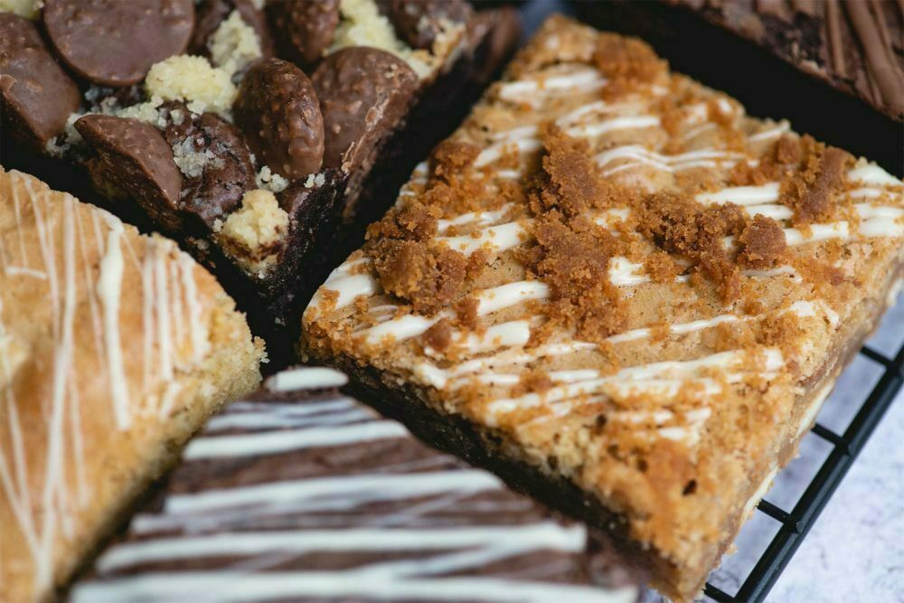 A close up image of a selection of Brownie and The Bean's chocolate brownies and blondies. All brownies are drizzled with white chocolate. Placed on a baking tray fresh out of the oven. The image is to promote our 3 month cake subscription.