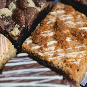 A close up image of a selection of Brownie and The Bean's chocolate brownies and blondies. All brownies are drizzled with white chocolate. Placed on a baking tray fresh out of the oven. The image is to promote our 3 month cake subscription.