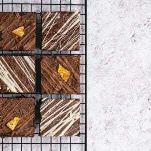 6 brownies without gluten on a baking grid. Three of these brownies are chocolate orange, which are drizzled with milk chocolate and have a candied orange on top and 3 brownies are Triple Chocolate which are a dark chocolate brownie drizzled with white chocolate. All 6 of the brownies are in a pack shot on a marbled background