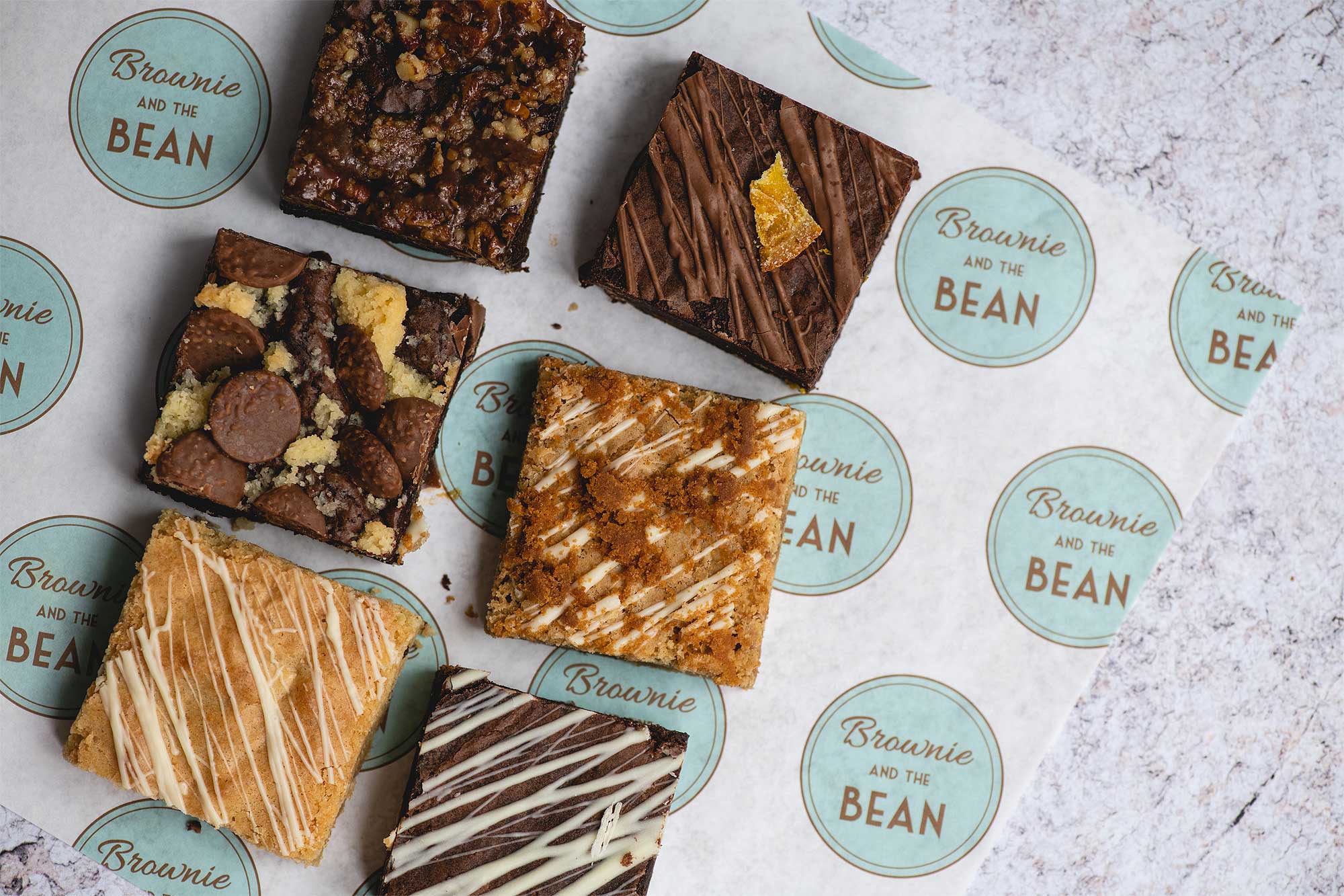 A laid out shot of our brownie deliveries by post. Photo contains our Mixed box of chocolate brownies on a printed greaseproof background