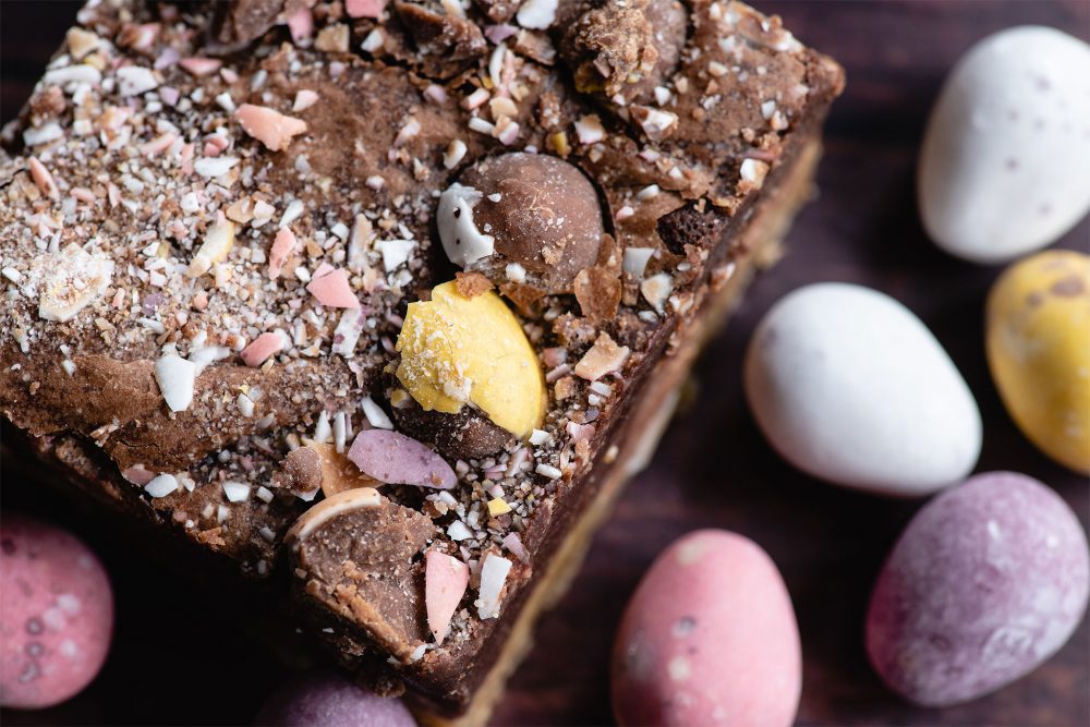 Mini Egg Brookie from our Easter Brownie Box. The the right are various pastel coloured chocolate Mini Eggs. To the left and centre of the image is a Brookie with crushed Mini Eggs on top.