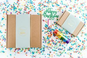 Birthday Party in a Box on a white background with confetti. There's two boxes, one square and a smaller rectangular box. There's also a confetti cannon, birthday topper and candle