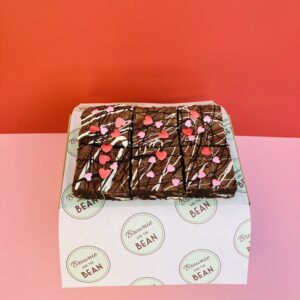 Valentine's Day brownies are no a branded sheet of Brownie and the Bean greaseproof paper with a pink and red background. The brownies have red and pink heart shaped sprinkles in different sizes on them