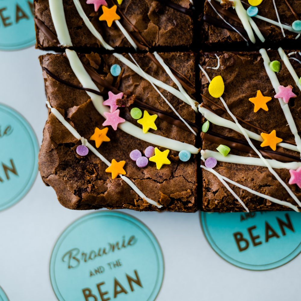 A close up of our birthday brownies for delivery. Triple Chocolate Brownies are drizzled in white chocolate and topped with Funfetti (which are bright coloured edible sprinkles). There are four in vision but the main focus is on the bottom left chocolate brownie.