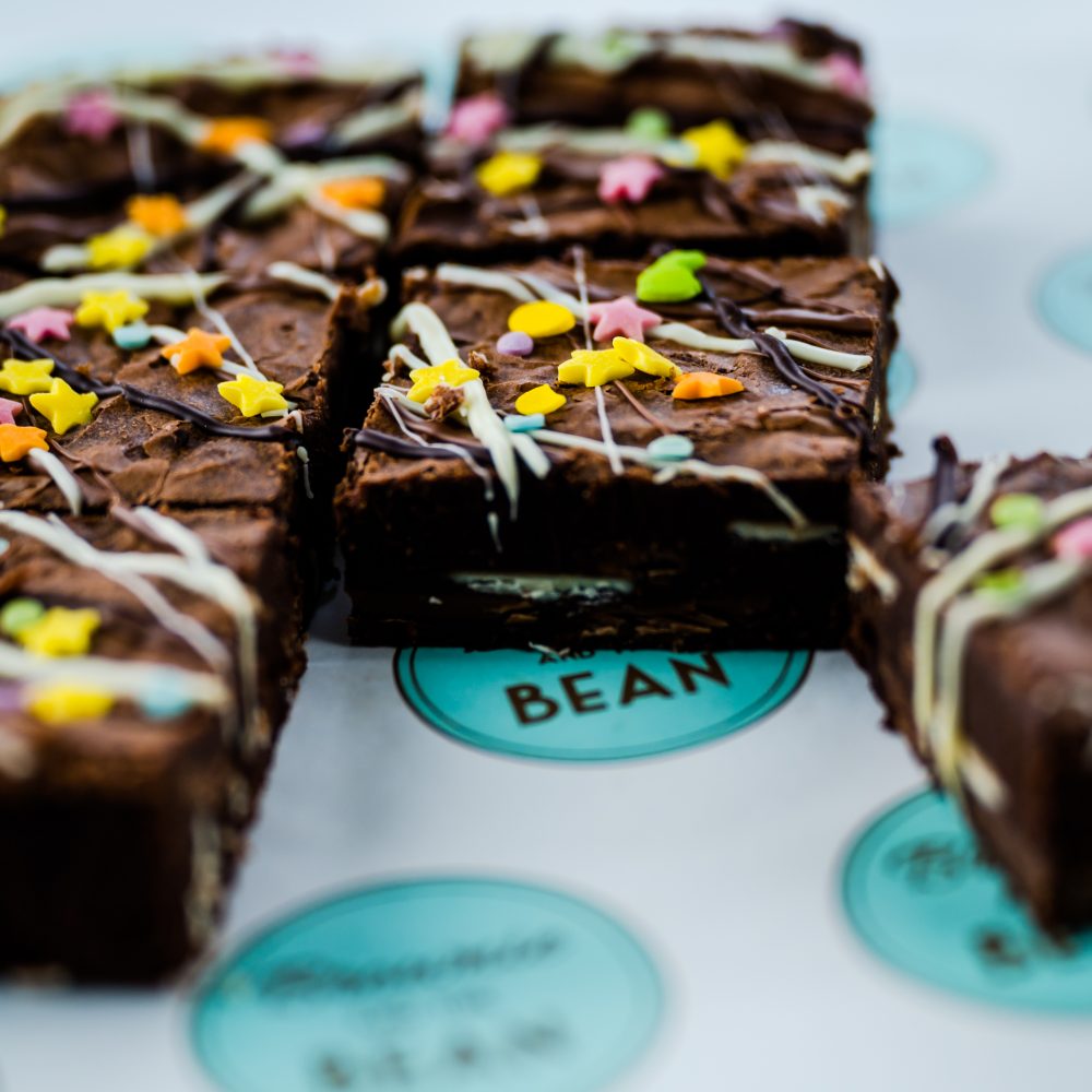 Triple Chocolate Funfetti Brownies with a white chocolate drizzle and brightly coloured sprinkles on top. There are six of them and one is moved to the side of the shot so you can see the profile of the chocolate brownie
