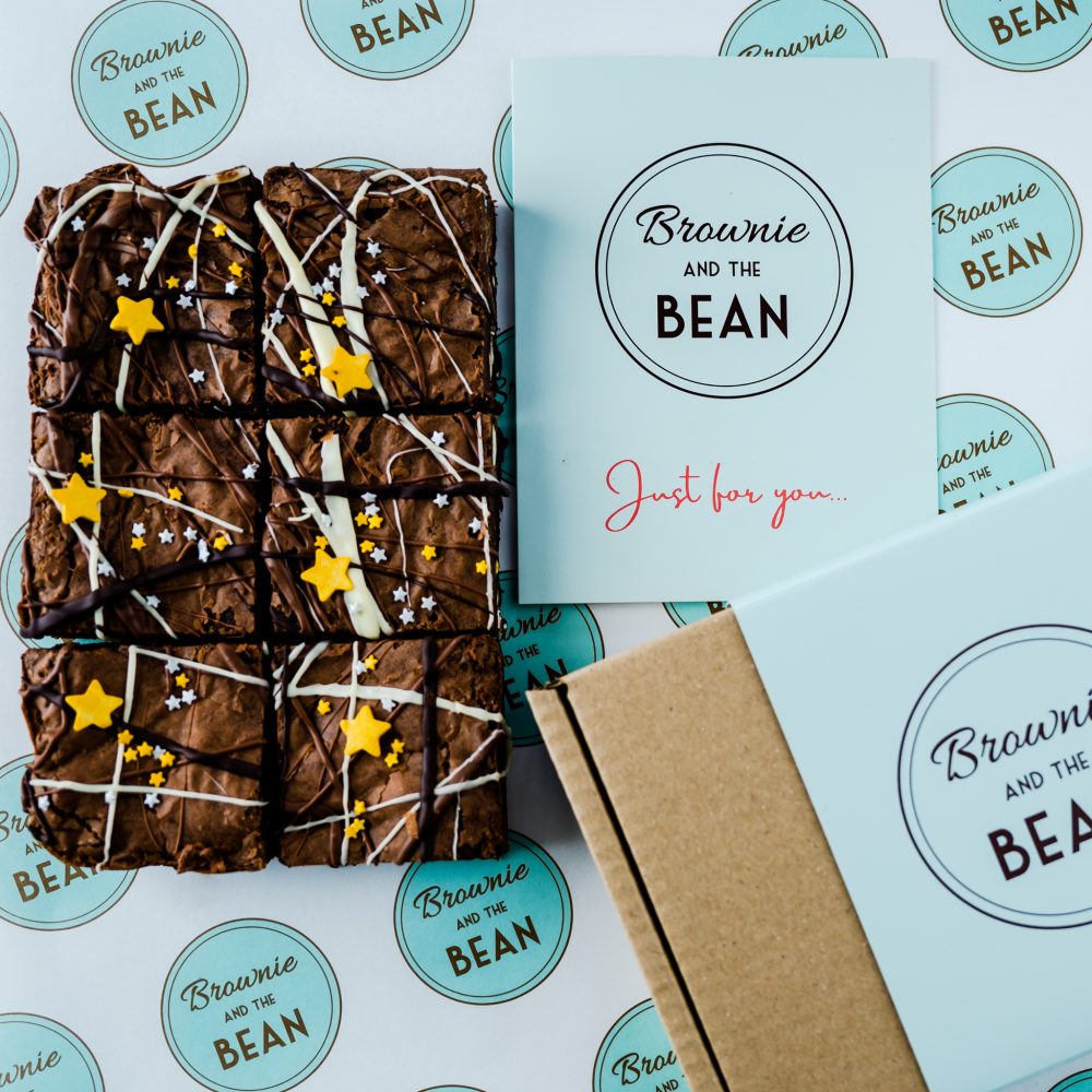 Triple Chocolate brownies drizzled with white chocolate and dark chocolate, topped with silver and gold star sprinkles from our Thank You Gift Set on a branded greaseproof background. There is a card and a Brownie and the Bean box also in vision