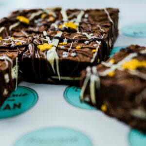 An image of bakery Brownie and The Bean's classic triple chocolate brownies. The images is advertising their “Thank You” Triple Chocolate Gift Set. There are 6 triple chocolate brownies in the photo that are all drizzled with white and dark chocolate. There are star shaped sprinkles of different sizes in gold and silver. The brownies are laid on branded paper.