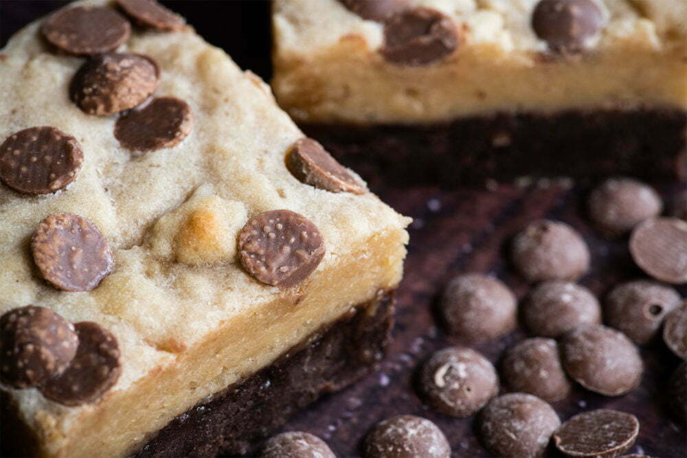 Our salted caramel cookie dough brownie is on a dark background. The top of the brownie is a cookie and light in colour. There are circular chocolate chips on top and sprinkled around the brownie. There is part of a salted caramel cookie dough brownie visible in the background.
