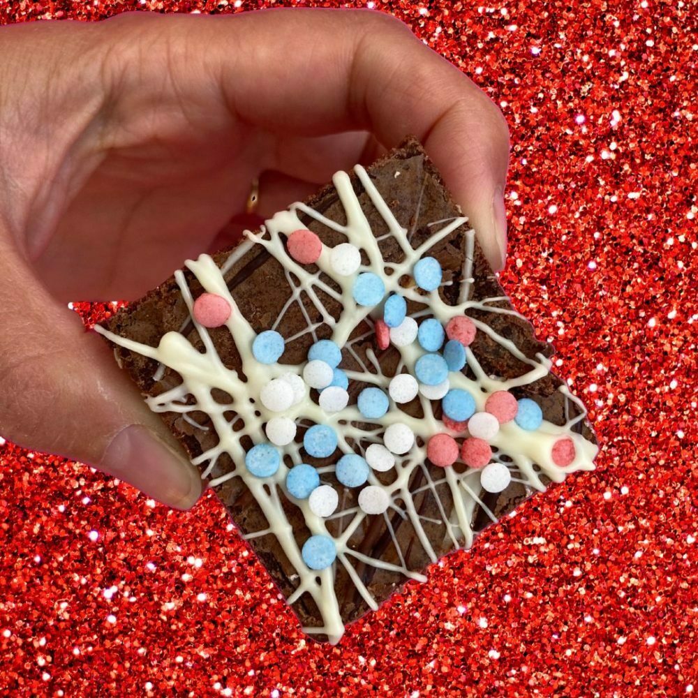 Chocolate Coronation Brownie is being held up by a human hand. The brownie is dark chocolate with a white chocolate drizzle. There are red, white and blue sprinkles on top. The background is red glitter.