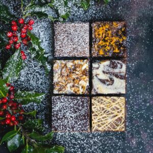 a variety of 6 different brownies on a navy background. There is holly on the left hand side. The background is navy and there is icing sugar resembling snow across the left of the image.