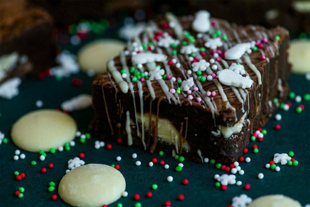 Christmas Triple Chocolate Brownie on a dark background with red, white and green sprinkles, white chocolate buttons and snowmen and snowflakes on the top.
