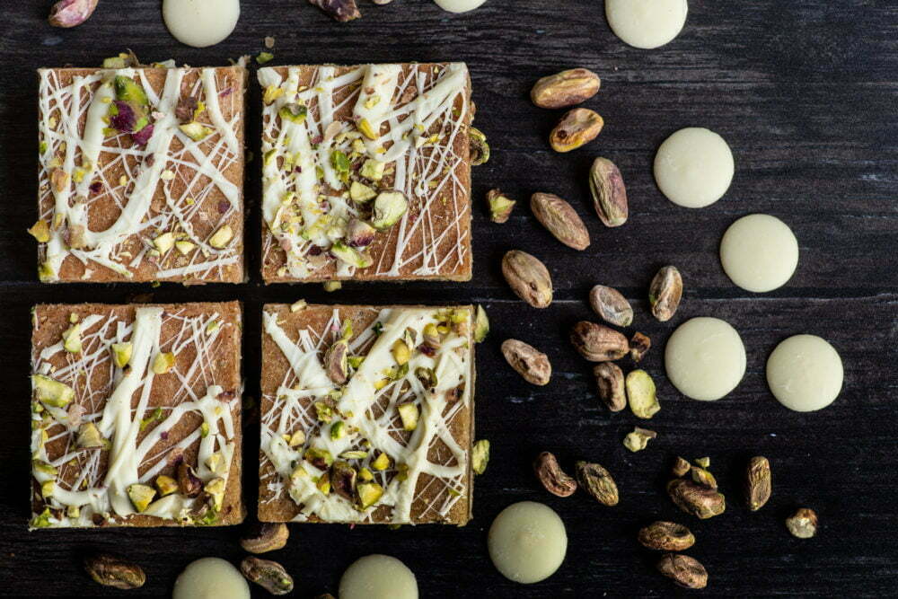 4 white chocolate and pistachio blondies on a dark background with additional white chocolate buttons and pistachios scattered across the page