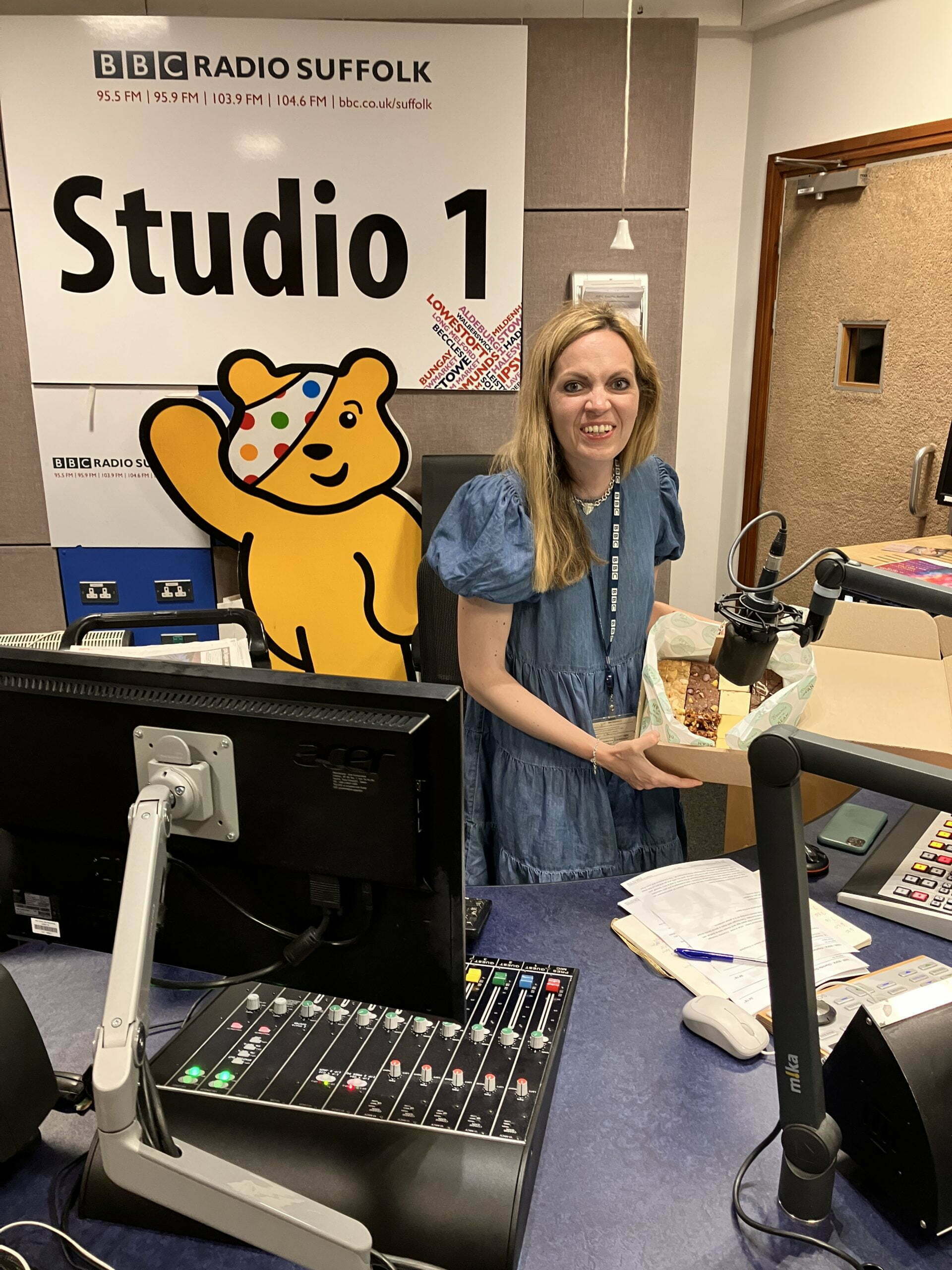 Pudsey Bear is waving in the background of Studio 1 at BBC Radio Suffolk. Sarah Lilley the presenter is behind the mixing desk with a large box of brownies. She's smiling.