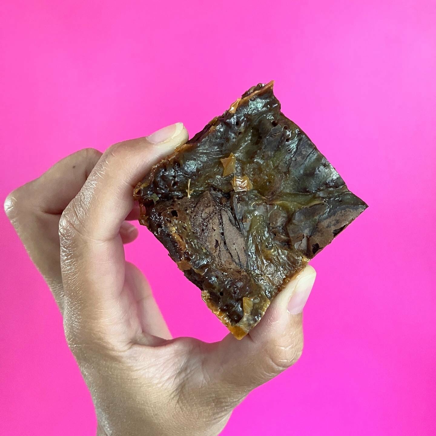 Malted Caramel Brownie being held up by a hand on a pink background.