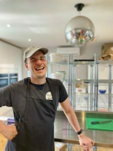Bean Daddy is stood in his branded t-shirt, wearing an apron in the bakery. There is a green chopping board behind him where he cuts his cake and there's a disco glitter ball hanging in the background.