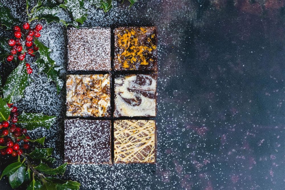 6 different varieties of brownie on a navy background with holly in the far left corner and a sprinkle of icing sugar, to give the effect of snow.