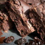 3 chocolate brownies stacked at a 45 degree angle with chocolate shards and raisins drizzled around. This is a close up shot with light bouncing off of the cracked Chcolate.