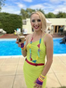A lady in 80's themed clothing is stood in front of a pool with a brownie ice cream sundae.