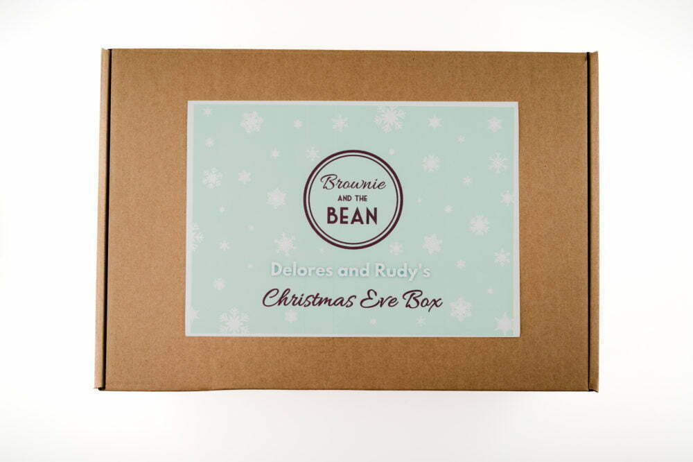 A brown Christmas Eve Box with personalised writing on it. The label is pale mint green with white snowflakes on. The Brownie and the Bean logo is in the middle (brown colouring), there is writing saying "Delores and Rudy's Christmas Eve Box".