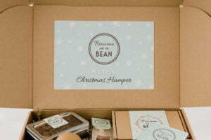 Our small Christmas Hamper is featured with a large pale mint green label with the brownie and the bean logo inside. In white writing the label says "Kate's: and below in scroll style writing it says "Christmas hamper". Within the hamper you can see hot chocolate stirrers, Christmas Cake in a tin, a box of brownies and a Christmas card.
