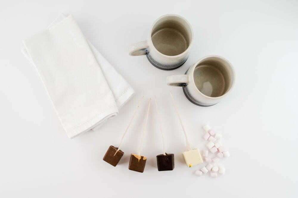 Two large mugs on a warm white background with two white napkins, 4 hot chocolate stirrers and marshmallows scattered.