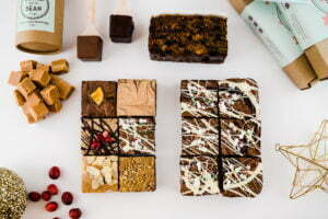 12 brownies laid out on a warm cream background. There are two Christmas baubles, 2 hot chocolate stirrers, a pile of fudge, a box of fudge, a sprinkling of cranberries and 2 brownie boxes layered on top of each other.