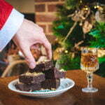 Father Christmas's hand is reaching for a brownie from a pile. There is a Christmas tree and a glass of sherry in the background.