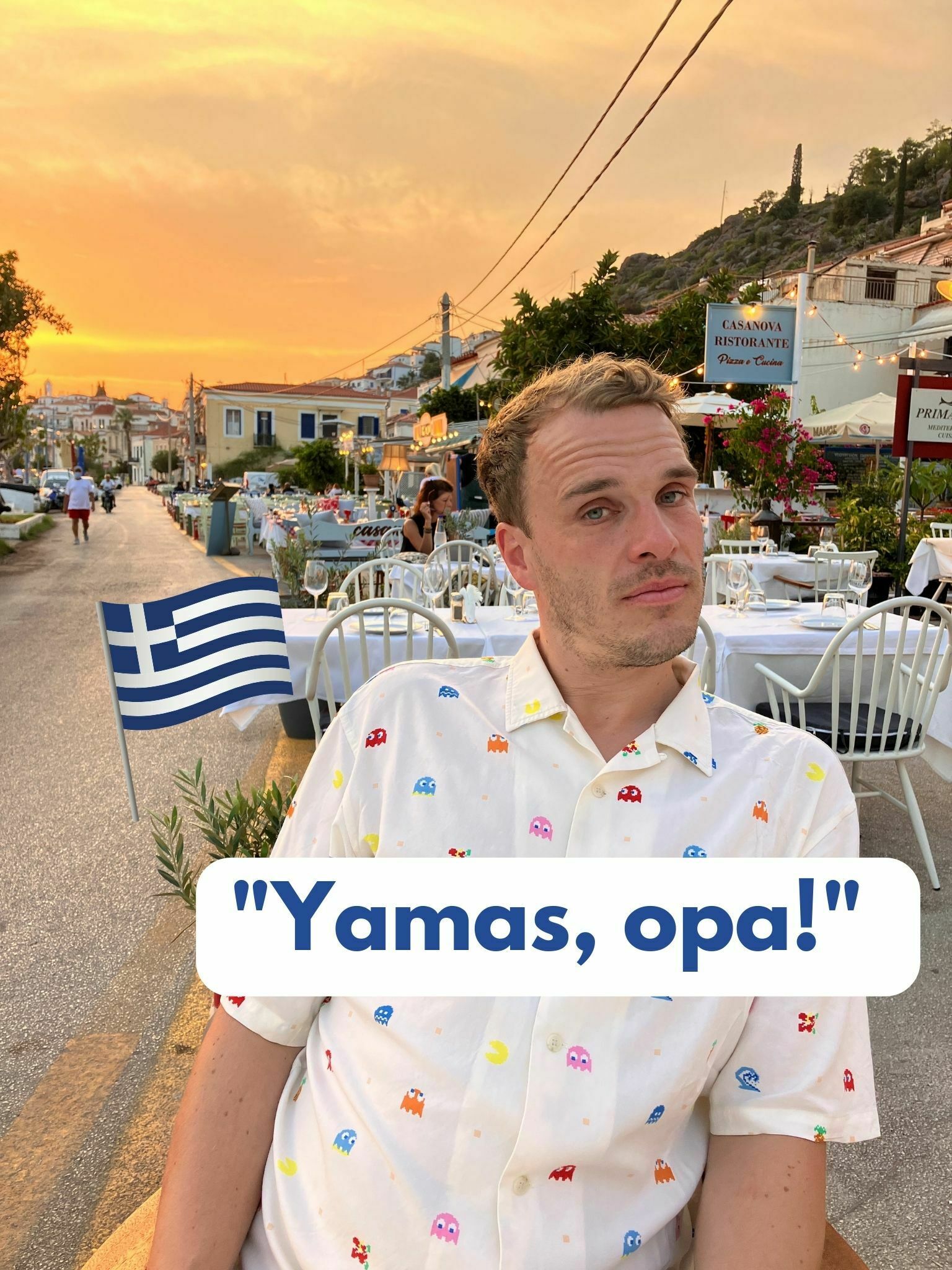 Text reads "Yamas, Opa" which means "cheers" in Greek. Bean Daddy has a strange look on his face. His in a decorative shirt with space invaders on it. There is a Greek flag to the left of him, a sunset in the background and a typically "greek" scene in the background.