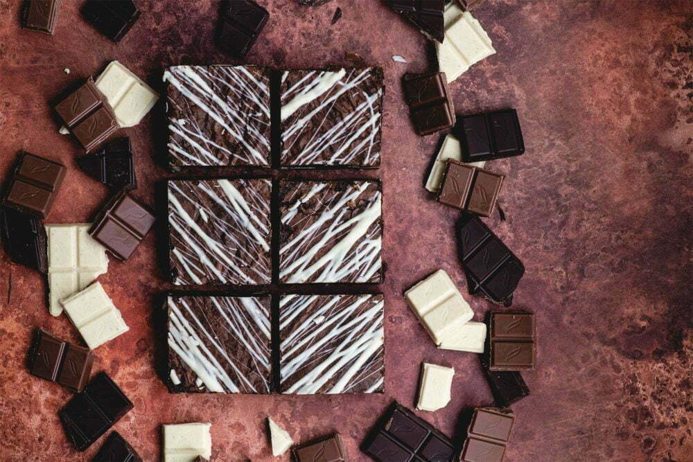 6 triple chocolate brownies on a dark burgundy background, with chunks of dark, white and milk chocolate surrounding it