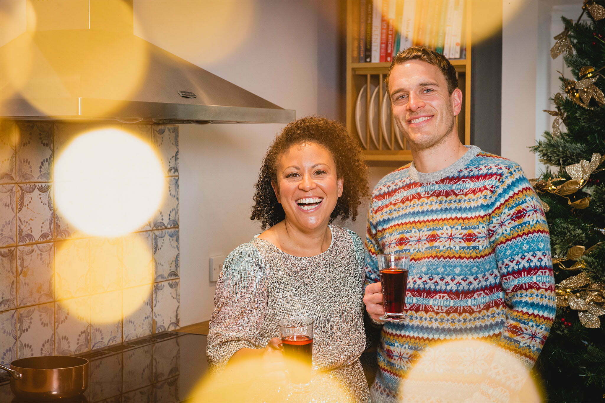 Luke and Charlotte are in their kitchen with a festive mise en scene. Luke is wearing a Christmas jumper and Charlotte is wearing a sequin dress. They're both holding drinks and are smiling.