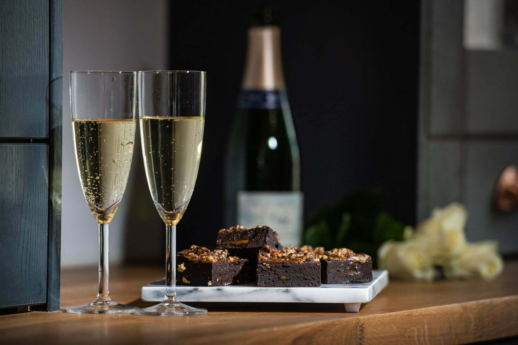 Brownies on a plate with two flutes of champagne and a bottle of champagne in the background. There are also white roses to the right of the bottle and brownies, indicating a wedding scene.