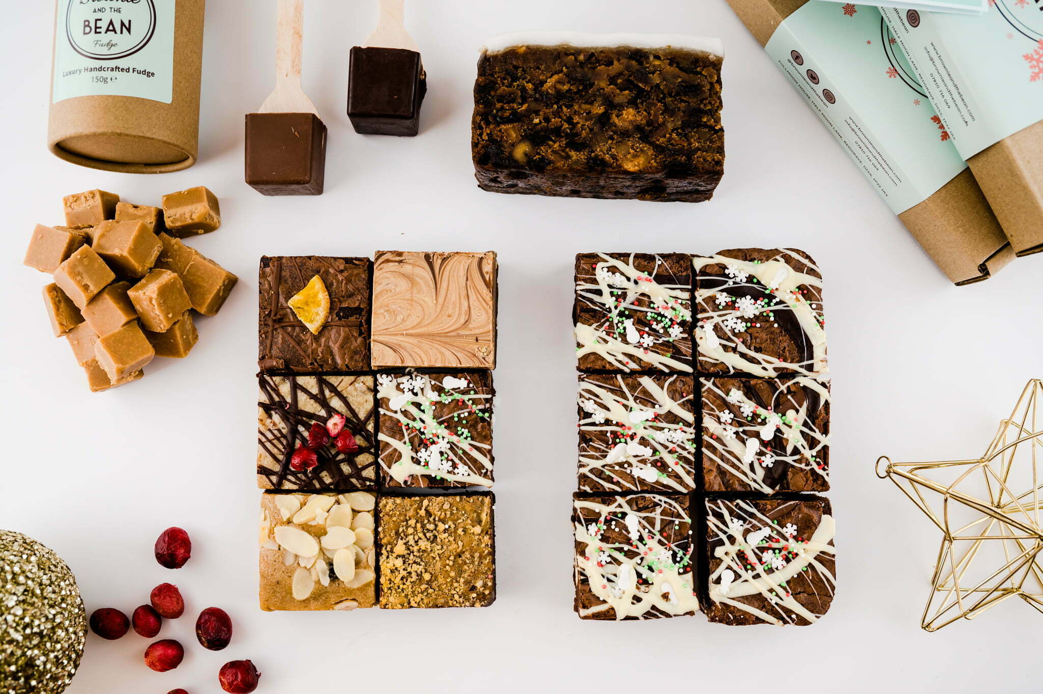 Christmas brownie hampers by post are laid out on a white background. There's cranberries, baubles, fudge, hot chocolate stirrers and Christmas Cake. There are also 2 brownie boxes and a box of fudge in the background.