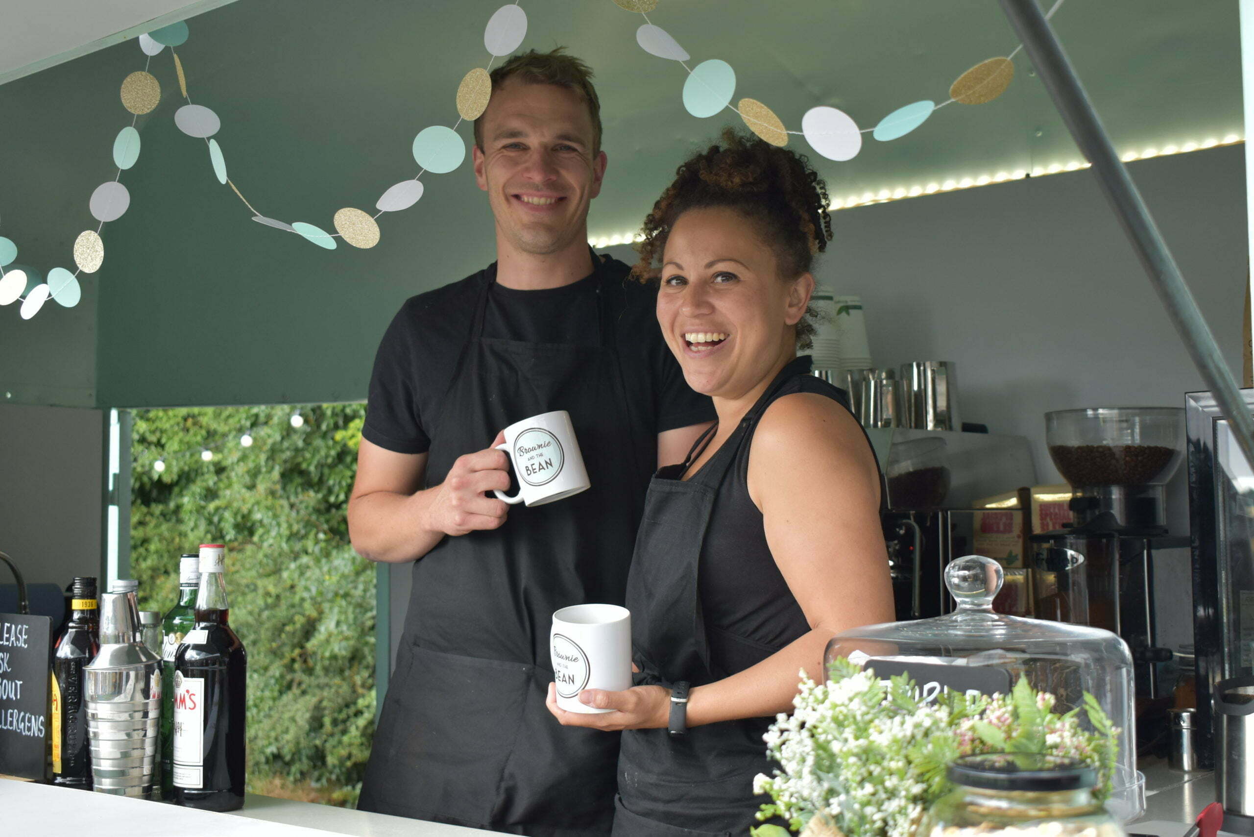 Charlotte and Luke ahead of starting their wholesale cake business, in their converted horse trailer. They're smiling and holding branded mugs. There is bunting in front of their faces and a cake dome to the right with foliage on it.