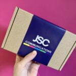 A brownie box is held up against a pink background. There is a brownie sleeve in Navy with white writing saying JSC Communications.