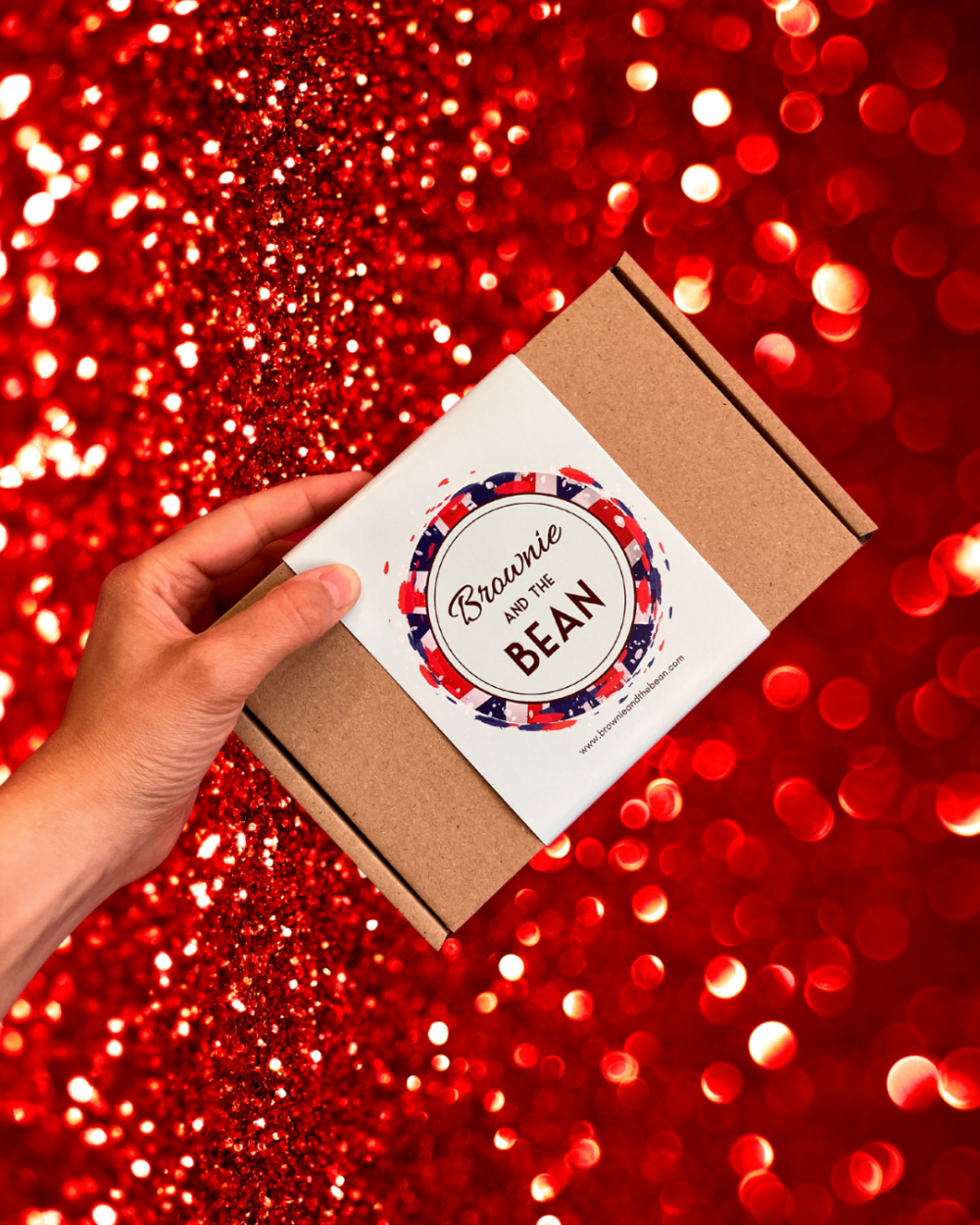 Our Best of British Brownie Box is being held up against a red glittered background. The brownie box has a Union Jack surrounding the Brownie and the Bean logo.