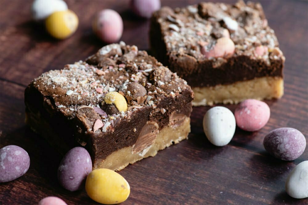 Our chocolate Mini Egg Brookie Easter brownies by post are on a brown background. There are two square shaped brownies with mini egg dust coated on them. There are mini eggs dotted around in pastel shades: yellow, white, pink and purple.