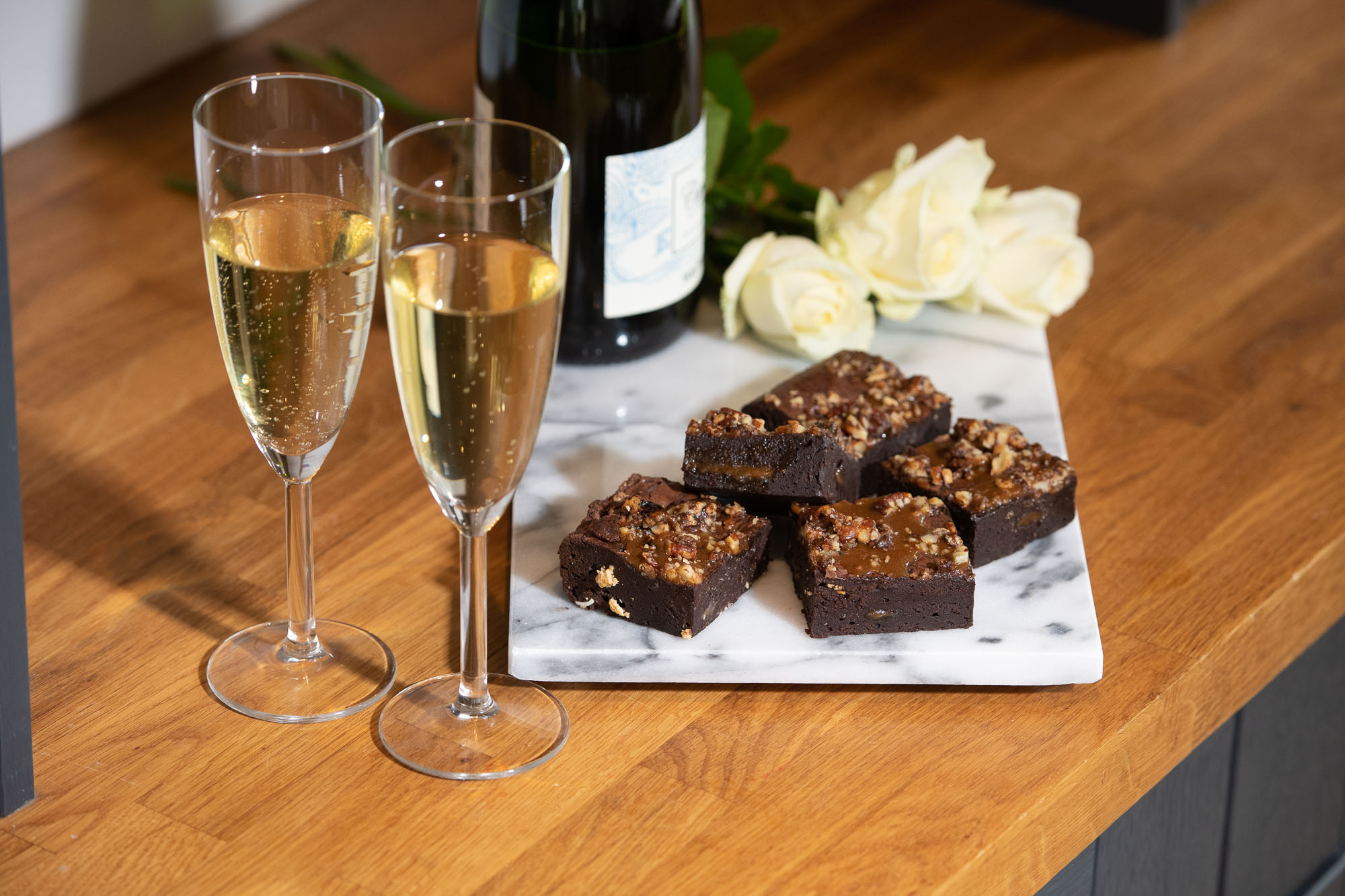 photo features mini wedding brownie stacks. The 5 stacked brownies are on a marble chopping board. The chopping board is on a wooden worktop. There are two champagne flutes in the foreground and there is a bottle of champagne and white roses in the background (on the marble) chopping board