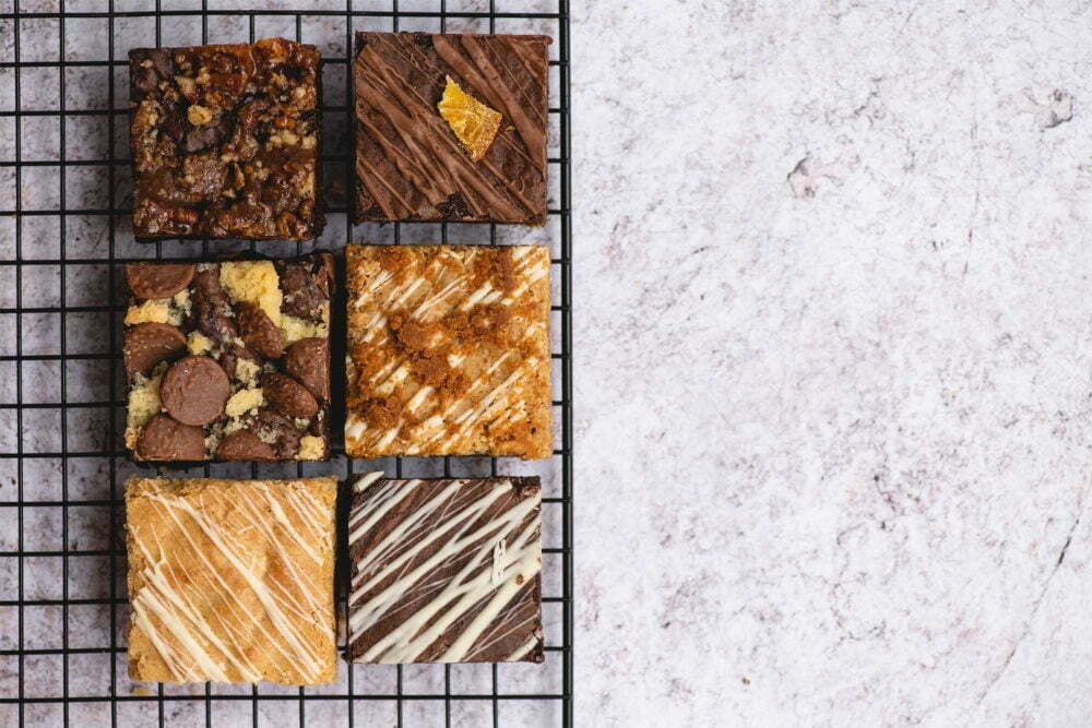 In the photo is our chocolate brownies by post. Our Chocolate Brownie Subscription is on a baking grid, laid on a marbled, light background. There are four chocolate brownies and two white chocolate brownies in the photo.