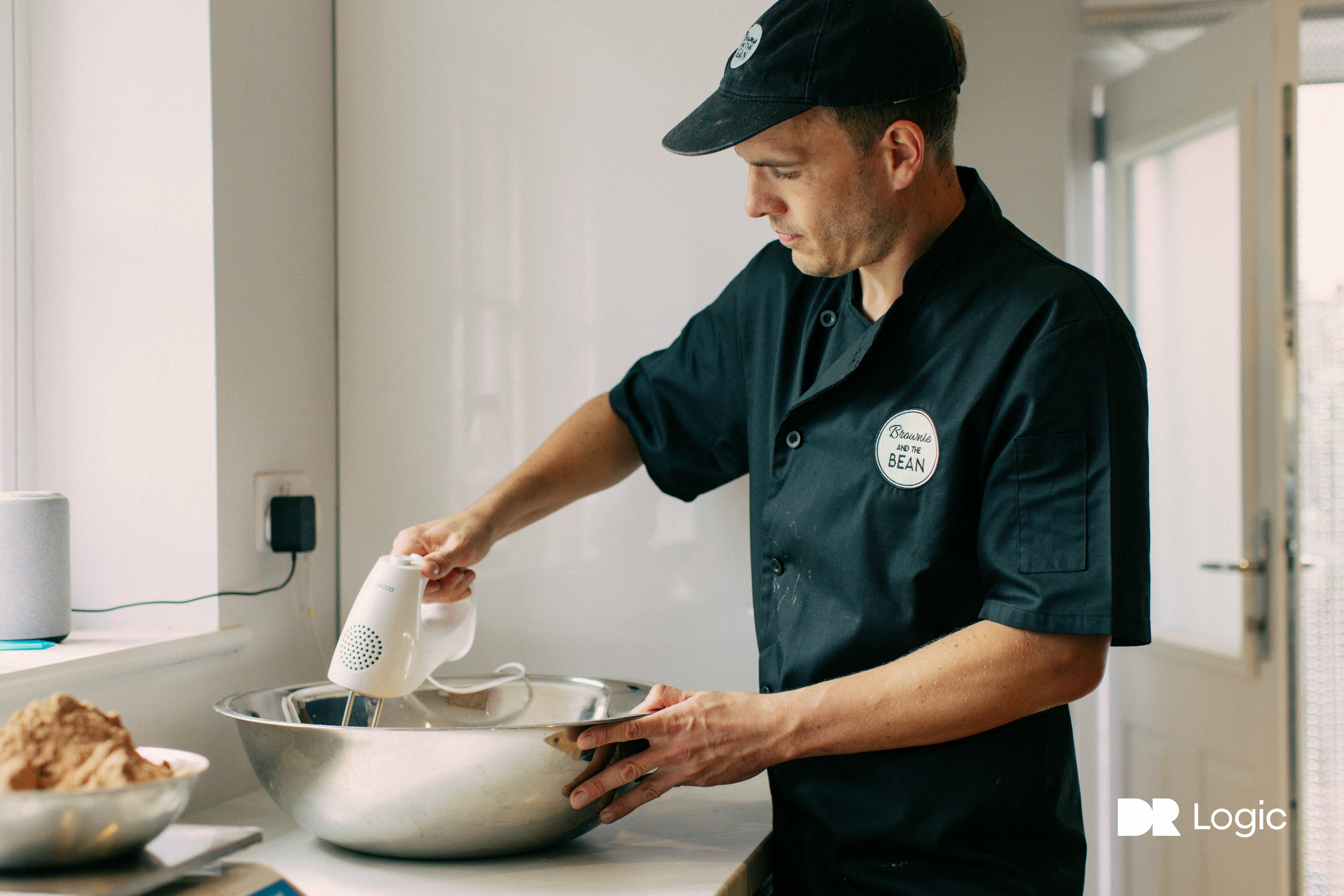 A close up of Bean Daddy in the Brownie and the Bean kitchen. He has a large silver mixing bowl and is using a handheld whisk. He is wearing black chef's whites with the Brownie and the Bean logo and he also has a black cap with the Brownie and the Bean logo on.