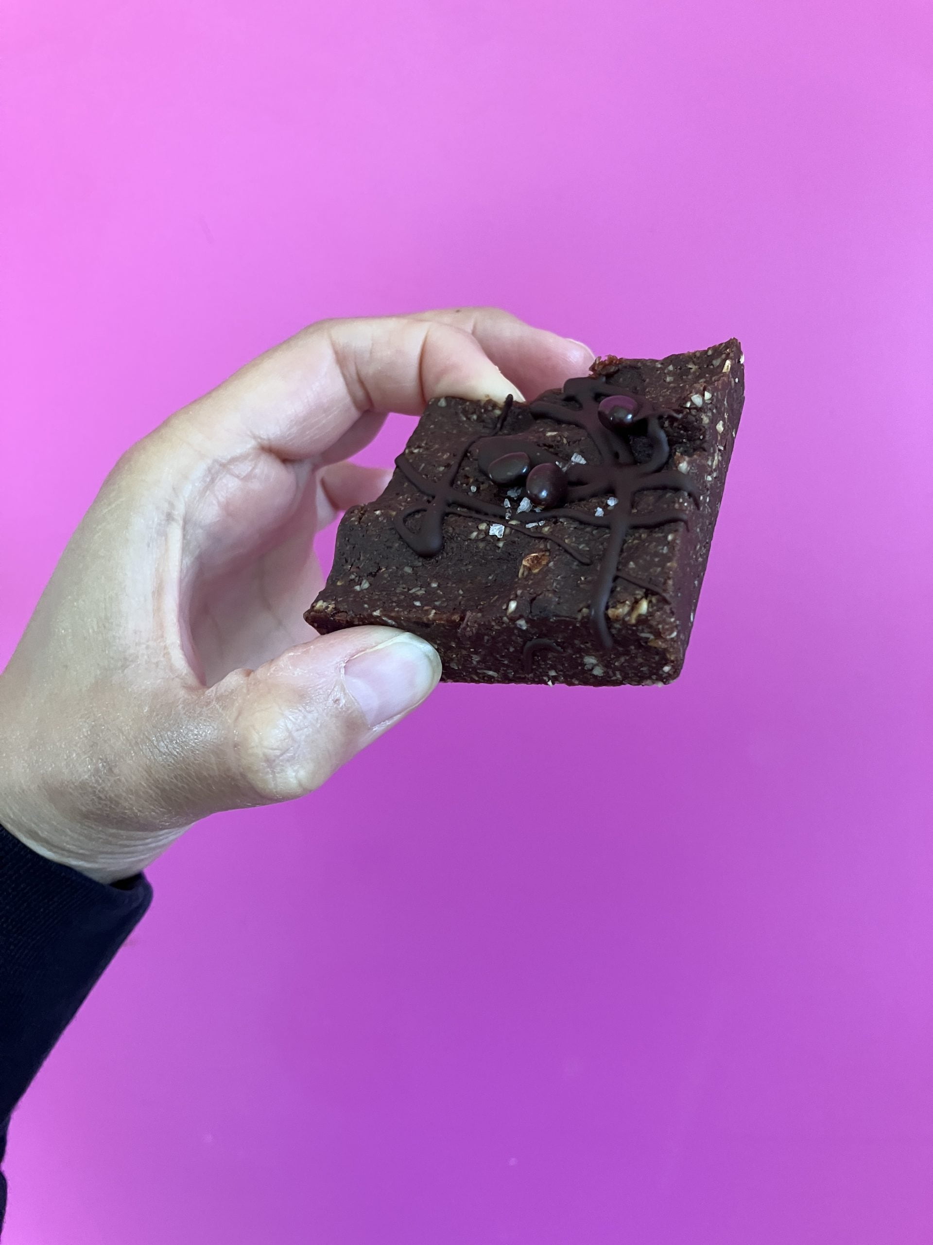 Charlotte (co founder) has her wrist in shot. She has a black top on which is slightly visible. She is holding one of their keto brownies in the air, against a pink background.