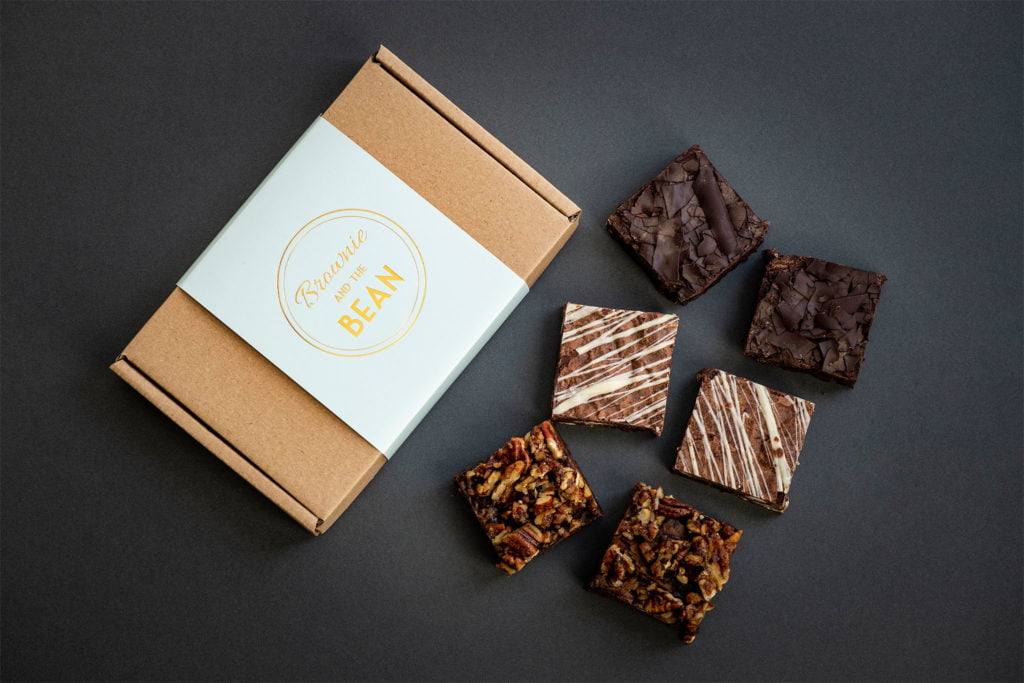 A brownie box is on dark background. The box is made of Kraft. The packaging sleeve is eau de nil and the Brownie and the Bean logo is in gold. In front of the box is 6 chocolate brownies. There are two of each flavour of the chocolate brownies.