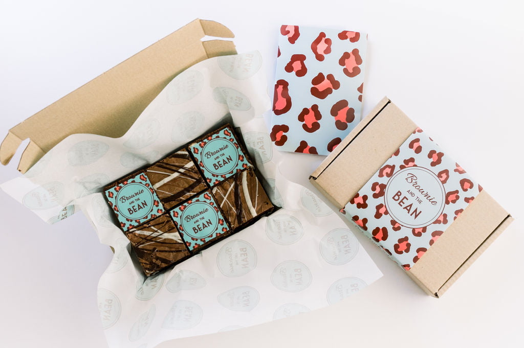 A sample of one of our corporate gifting solutions is in the photograph. The background is white. There is a Kraft box of brownies to the right. The outer sleeve is leopard print and turquoise and pink. The Brownie and the Bean logo is in the centre of the packaging. Above and to the left is a greetings card with turquoise and pink leopard print packaging. Further to the left is an open box of chocolate brownies. Three of them have the Brownie and the Bean logo in Turquoise and pink leopard print. The other three chocolate brownies have a white chocolate drizzle on the top.