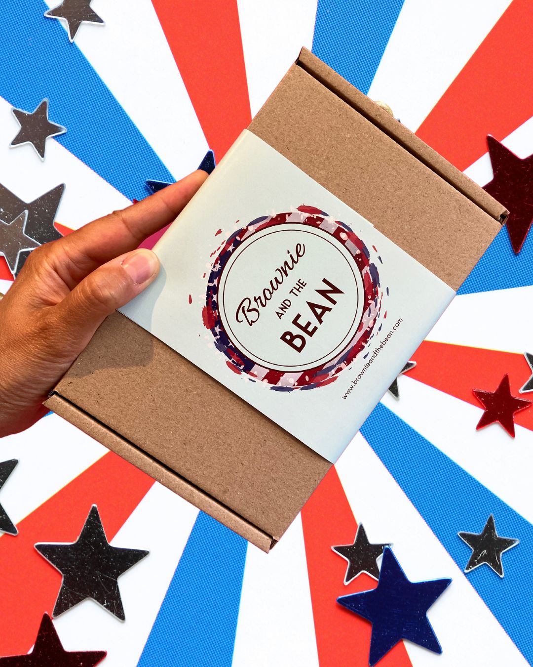 American themed background with red, white and blue strips emerging from the centre of the photo. There are stars placed sporadically around. In the centre of the image is a hand holiday an American themed brownie box. The box is Kraft, the strap is eau de nil and there is an American flag in the shape of a circle, behind the eau de nil Brownie and the Bean logo.