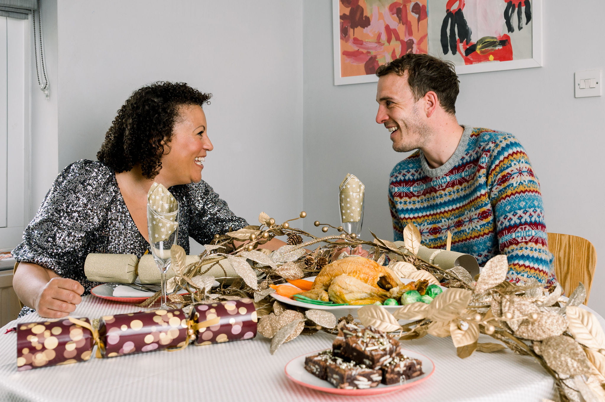 Charlotte and Luke (founders of Brownie and the Bean) are sat at a Christmas laid table. They are gazing lovingly into each other's eyes. There is a full Christmas spread of food including a roasted chicken and seasonal vegetables. There is a pile of Christmas brownies placed at the front. This photo forms as part of their Christmas in July 2023 campaign.