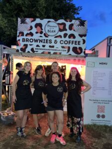 Brownie and the Bean at Latitude Festival. There is a sign above head hight with the Brownie and the Bean logo and "Brownies and Coffee" written in bold type font. There are 4 women and one man in the photo. These are the Brownie and the Bean staff. They're all wearing black with a gold Brownie and the Bean logo on. Everyone is smiling. There is a menu to the right hand side.