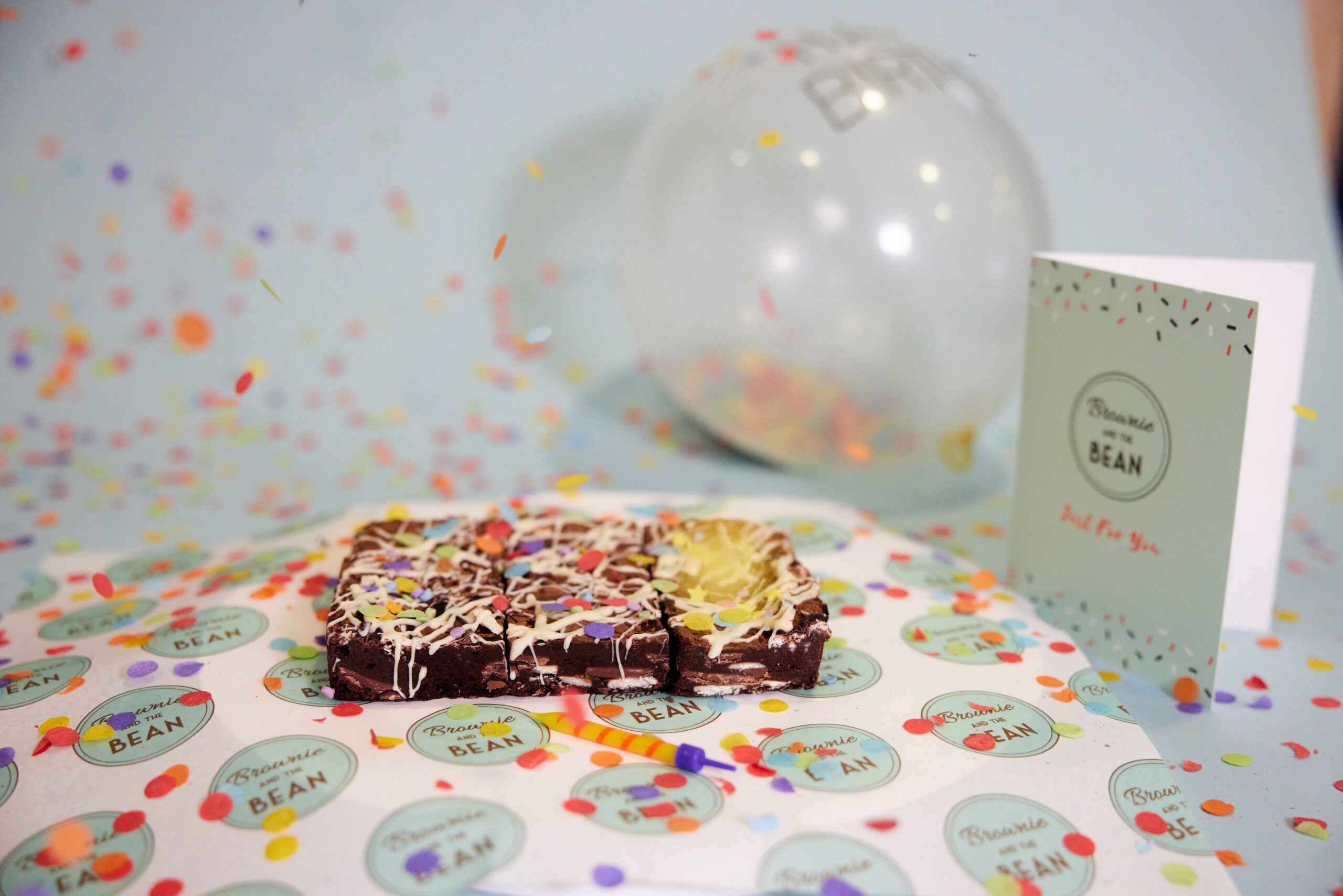 A scene of birthday brownies on branded Brownie and the Bean greaseproof paper, a ballon, a branded Brownie and the Bean card and bright coloured confetti flying in the air.