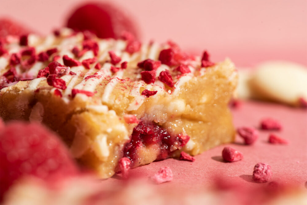 Raspberry Eton Mess Blondie is on a pink background with raspberries and white chocolate scattered around