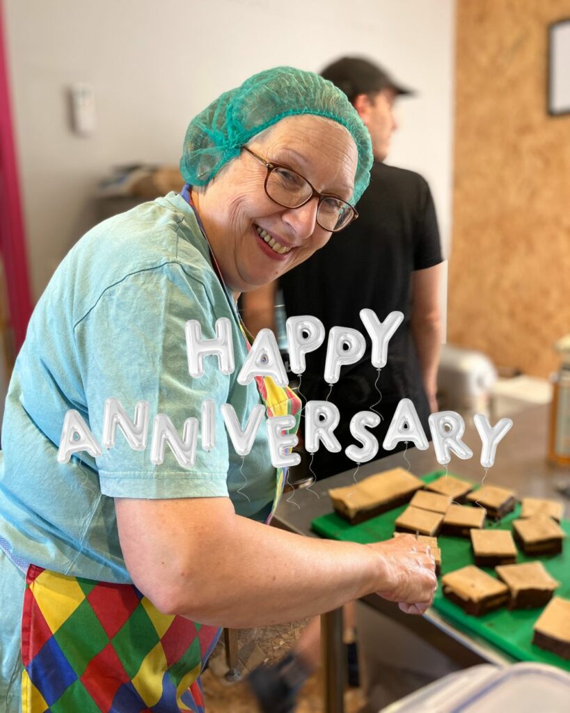 Super Sue smiling whilst chopping brownies. The title reads Happy Anniversary in bubbly balloon style writing.