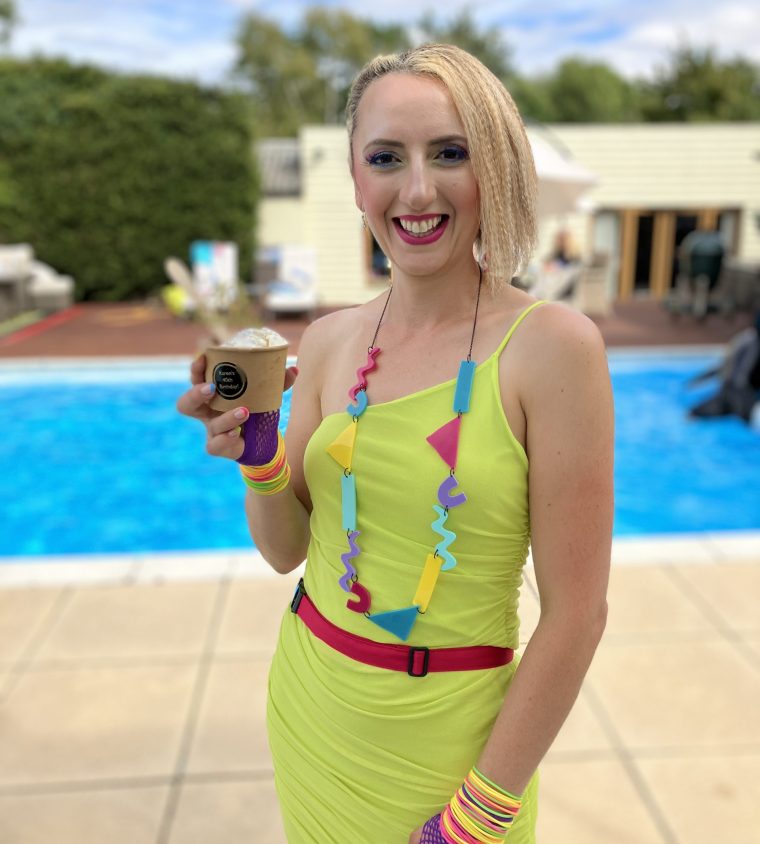 A lady in 80's themed clothing is stood in front of a pool with a brownie ice cream sundae.